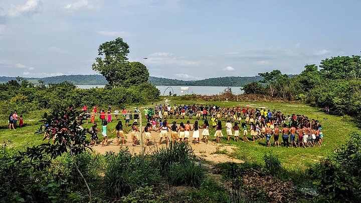 Eine Gruppe Munduruku hält eine Zeremonie ab und tanzt im Kreis im Amazonasregenwald. Im Hintergrund ein Fluss