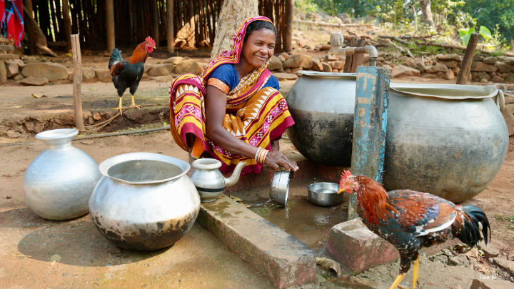 eine indische Frau sitzt lächelnd an einer Wasserstelle und spült, neben ihr sind wassergefäße und zwei Hähne