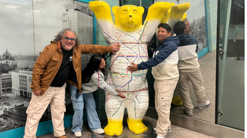 Der Berliner Bar als Figur mit dem U-Bahnnetz auf dem bauch hebt die Hande. Drei Brasilianner:innen stehen um ihn herum