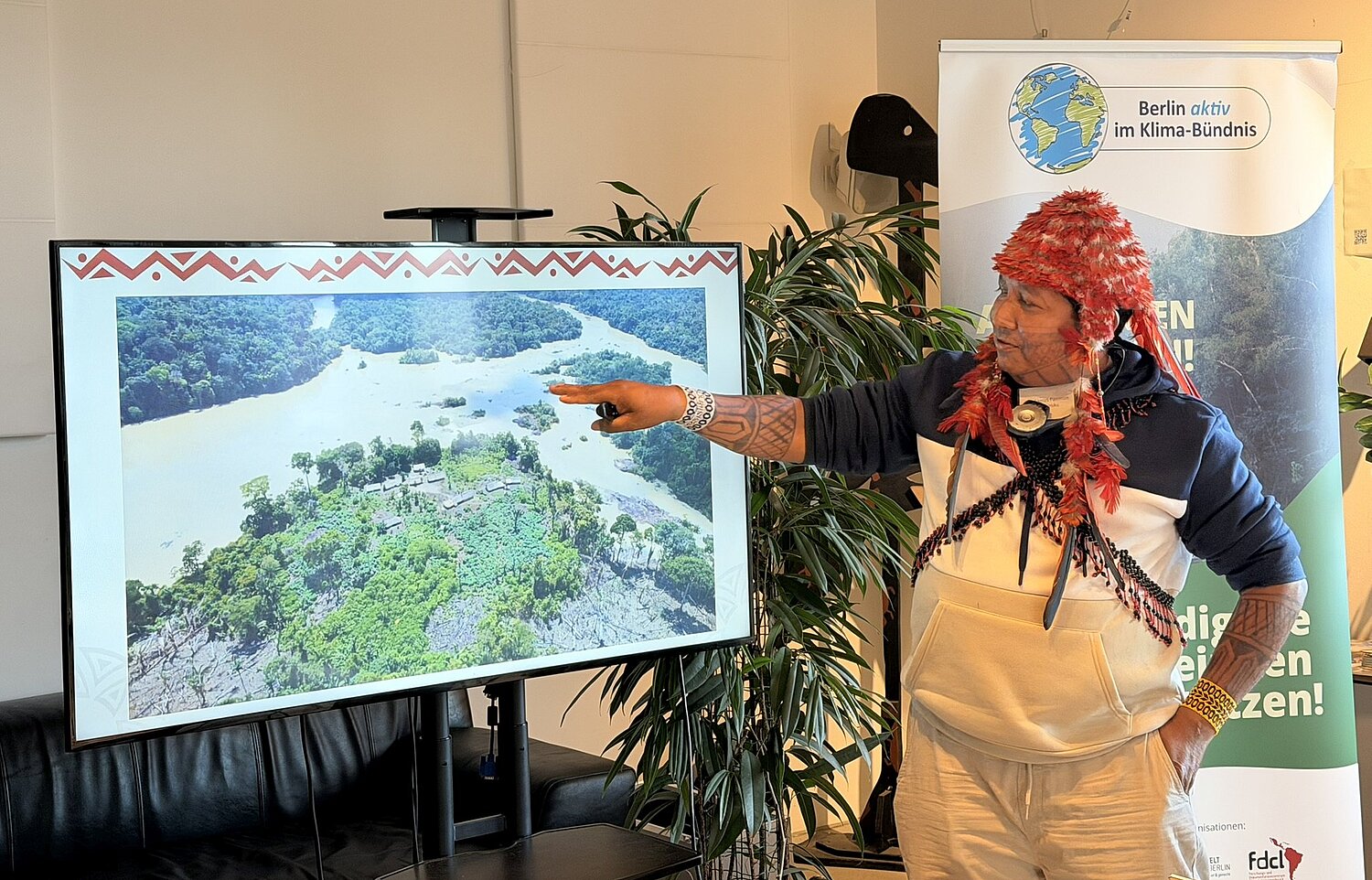 Ein Mann in der traditionellen Kleidung der Munduruku hält eine Präsentation. Im Hintergrund ein Banner der Initiative Berlin aktiv im Klimabündnis