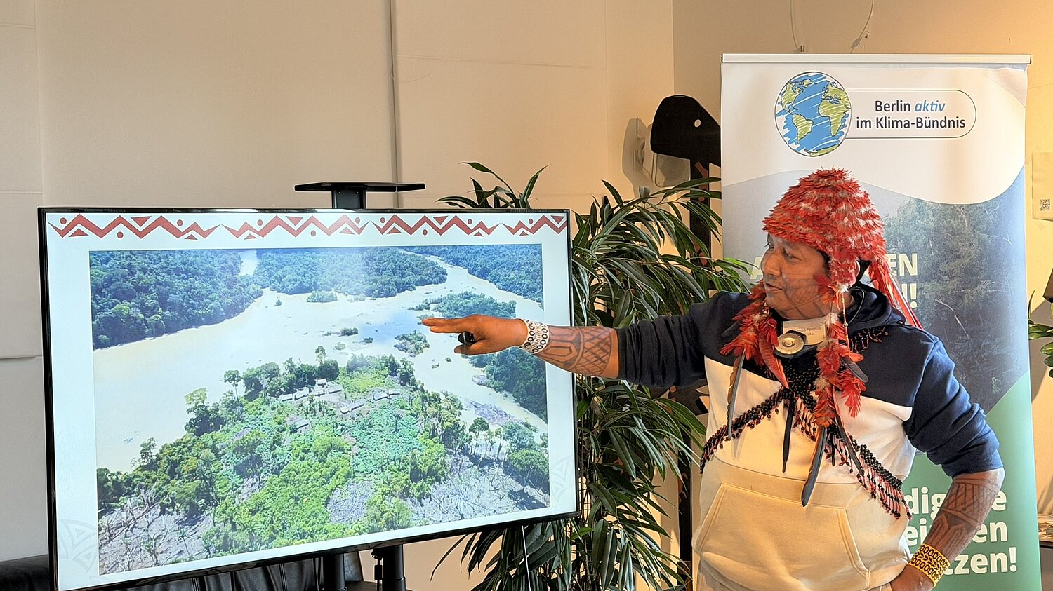Ein Mann in der traditionellen Kleidung der Munduruku hält eine Präsentation. Im Hintergrund ein Banner der Initiative Berlin aktiv im Klimabündnis