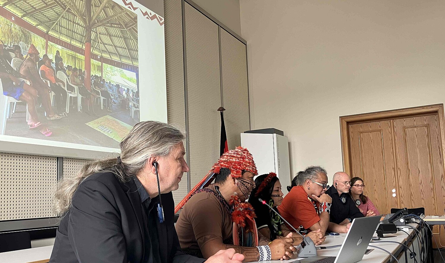 8 Personen sitzen in einer Reihe an einem Tisch mit Mikros. Im Hintergrund ein Foto einer Versammlung im Amazonasregenwald