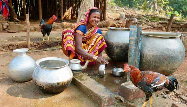 eine indische Frau spült lächelnd an einer Wasserstelle. Neben hier stehen Wasserbehälter und zwei Hähne laufen herum