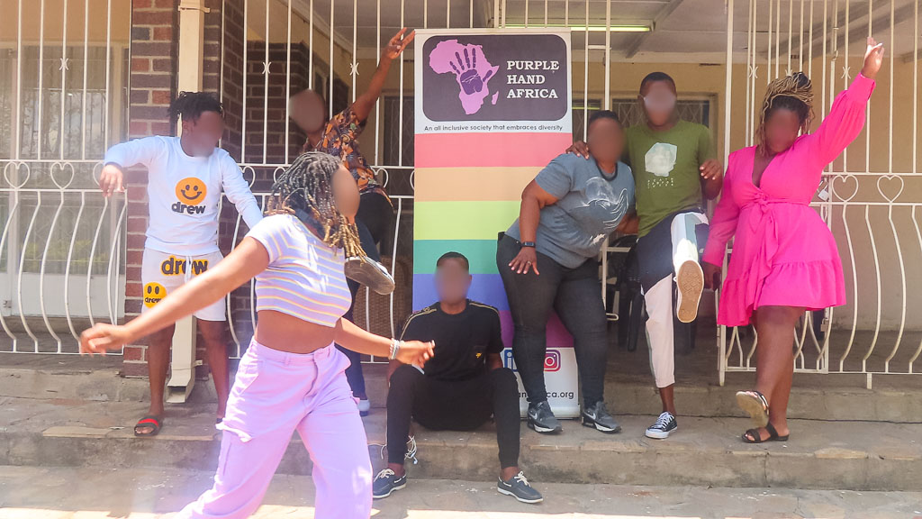 6 queere Personen stehen vor einem Haus mit dem Logo von Purple Hand