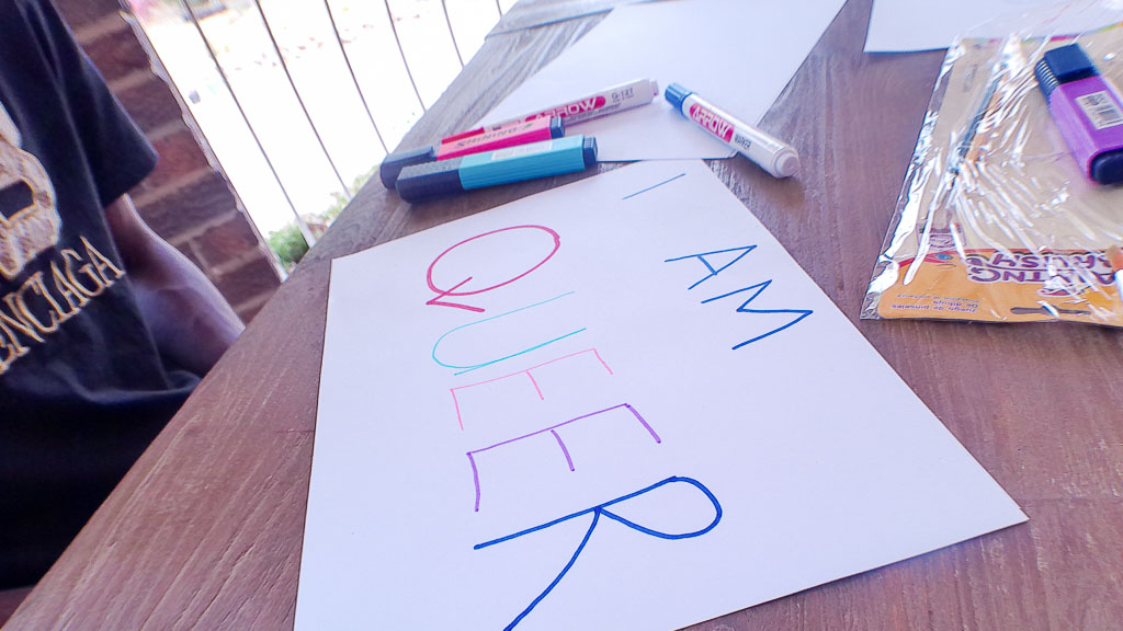 Eine Person sitzt an einem Tisch, vor ihr ein Blatt mit dem selbstgeschriebenen bunten Schriftzug "I am queer"